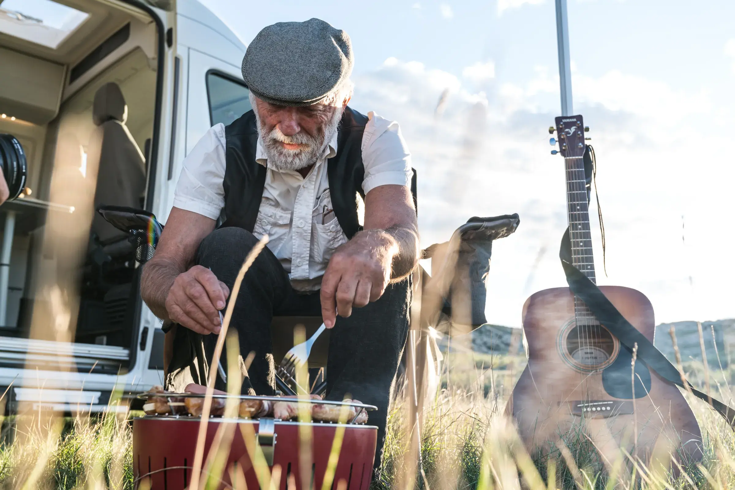 Mann som lager mat på bål ved bilen, gitar lent mot karosseriet