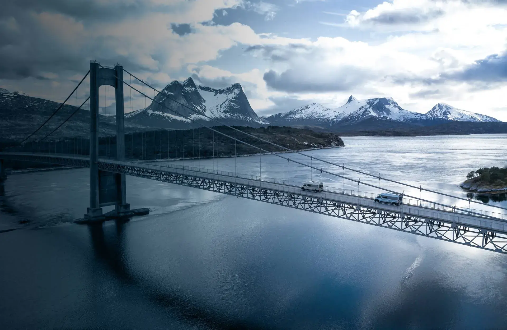 Tourne bybobil på hengebro over fjord, med snødekte fjell i bakgrunnen
